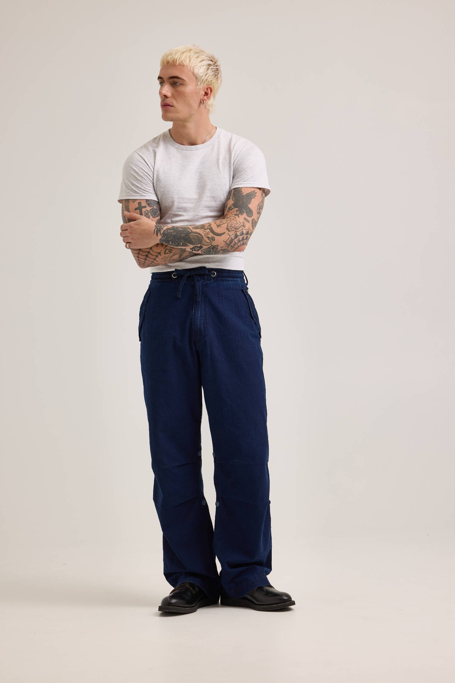 A person with blonde hair and tattoos stands wearing the crisp white Alpha X Lee Snowpant shirt and versatile blue pants made from ripstop cotton by Alpha Industries.