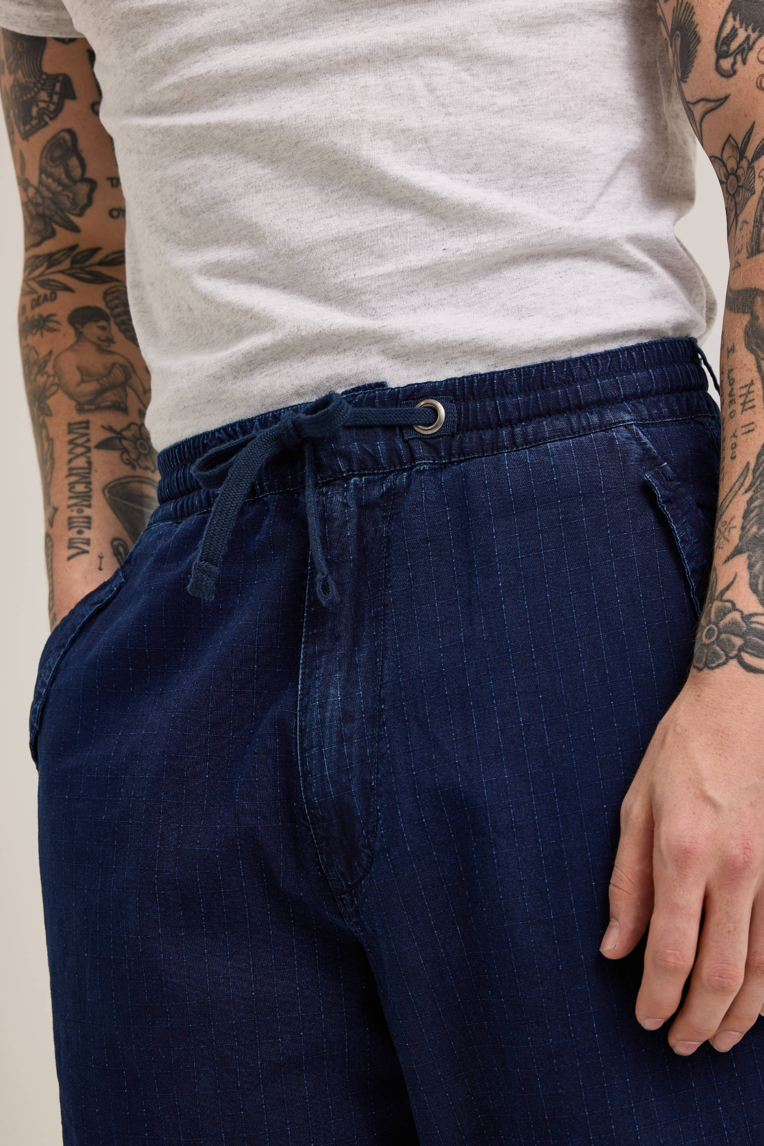 The person is wearing a white t-shirt and the versatile Alpha X Lee Snowpant by Alpha Industries, blue and pinstriped with a drawstring, showcasing their arm tattoos.