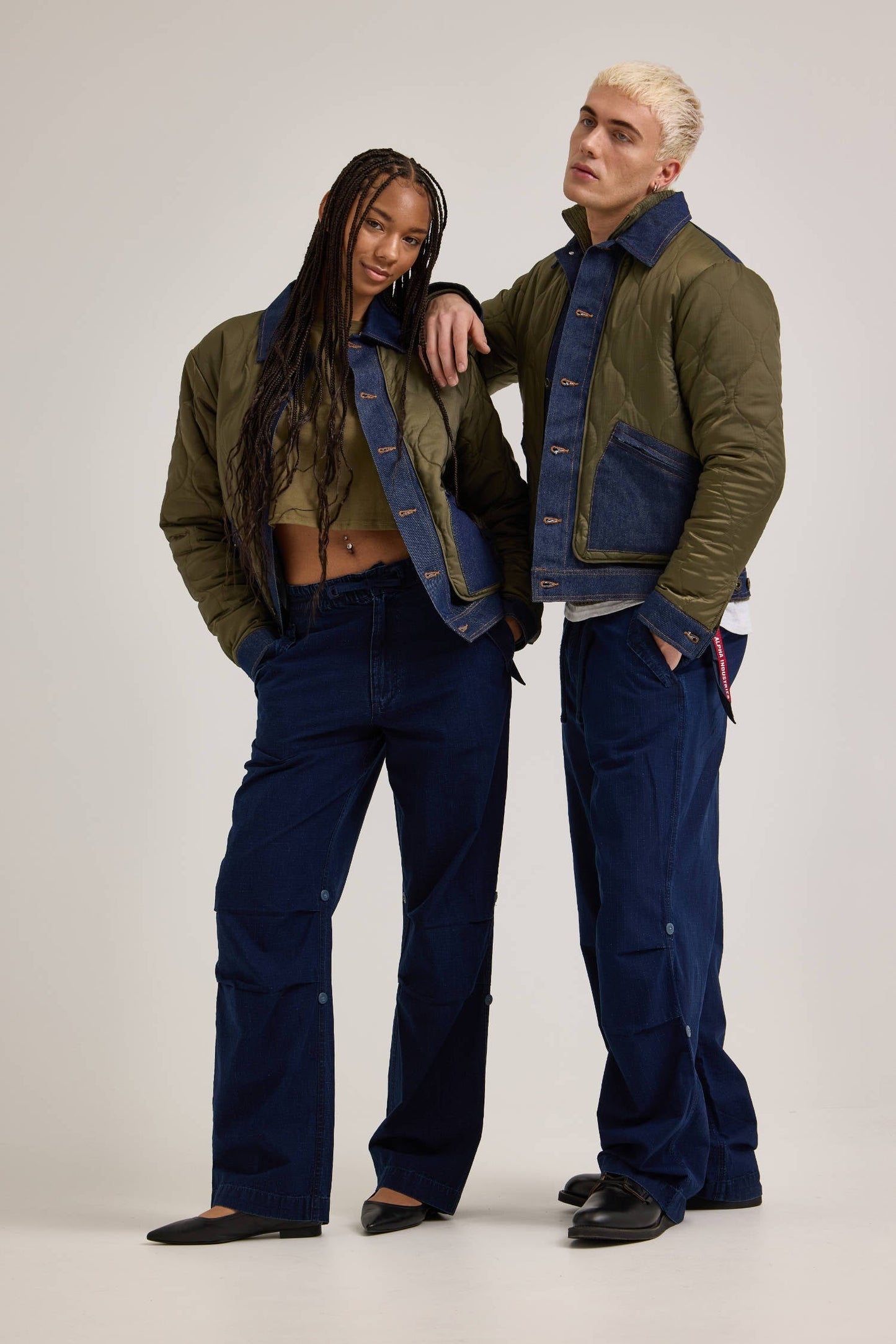 Two individuals stand side by side, wearing denim and olive green jackets. They are both outfitted in Alpha Industries / Lee Alpha X Lee Snowpant in indigo and black shoes, maintaining neutral expressions.