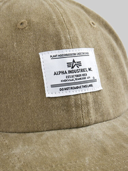 A close-up of the vintage-inspired olive Battlewash Cap by Alpha Industries, featuring a white label with company details and a caution not to remove it.