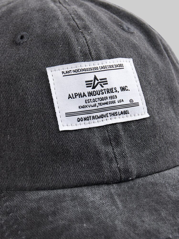 Close-up of the Battlewash Cap, a vintage-inspired gray baseball cap featuring a white label with text, including Alpha Industries and other details in black font.