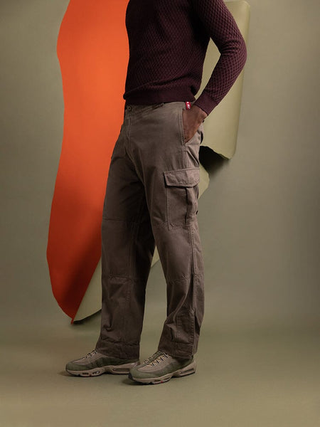 A person stands to the side, wearing the Belgian M-64 Pant in brown by Alpha Industries.