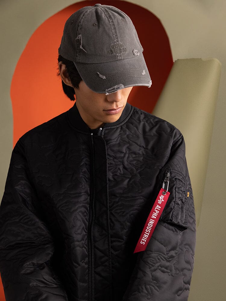 An individual wearing an Alpha Industries Camo Quilted MA-1 Bomber Jacket in black featuring a red tag on the sleeve and a utility pocket.