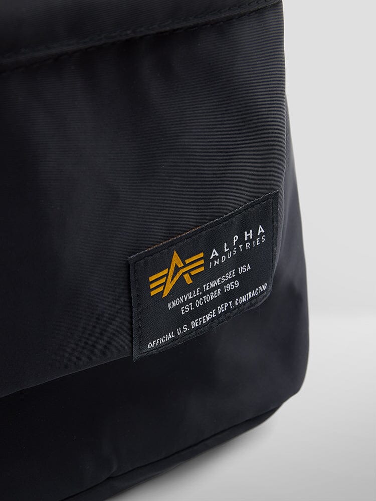 Close-up of the Crew Backpack by Alpha Industries in black flight nylon, featuring an Alpha Industries label with the company logo and establishment details.