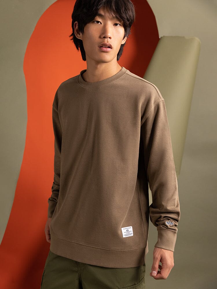 A person wearing the Essential Crewneck in coyote brown from Alpha Industries..