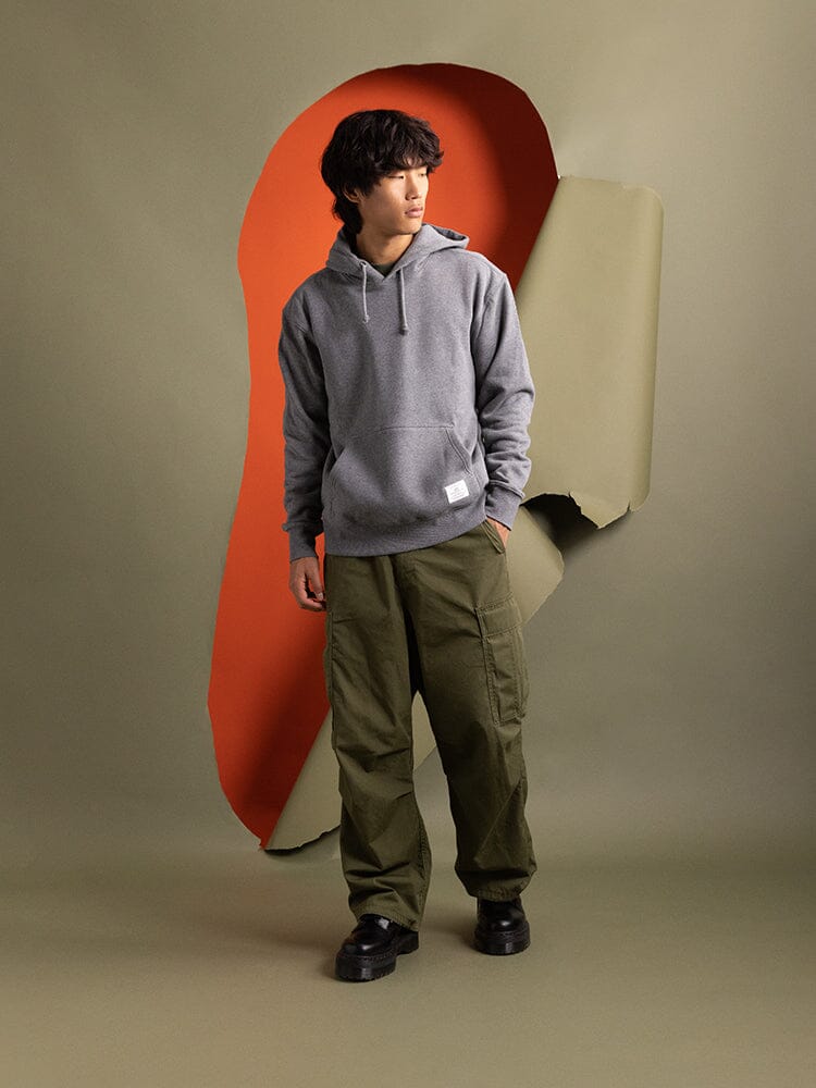 A person wearing the Alpha Industries Essential Hoodie in medium charcoal heather and green pants poses.