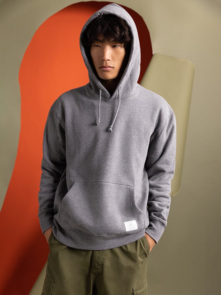 A person stand with his hands in his pockets, wearing a medium charcoal heather Essential Hoodie from Alpha Industries.