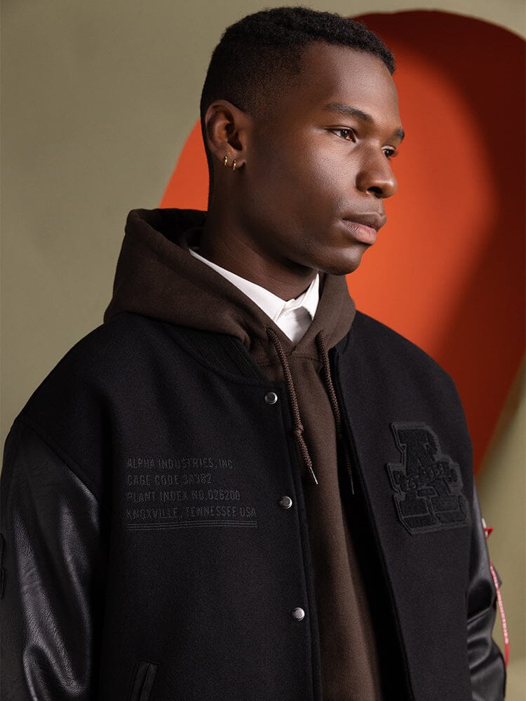 A person wearing the Faux Leather MA-1 Varsity Bomber Jacket in black by Alpha Industries.