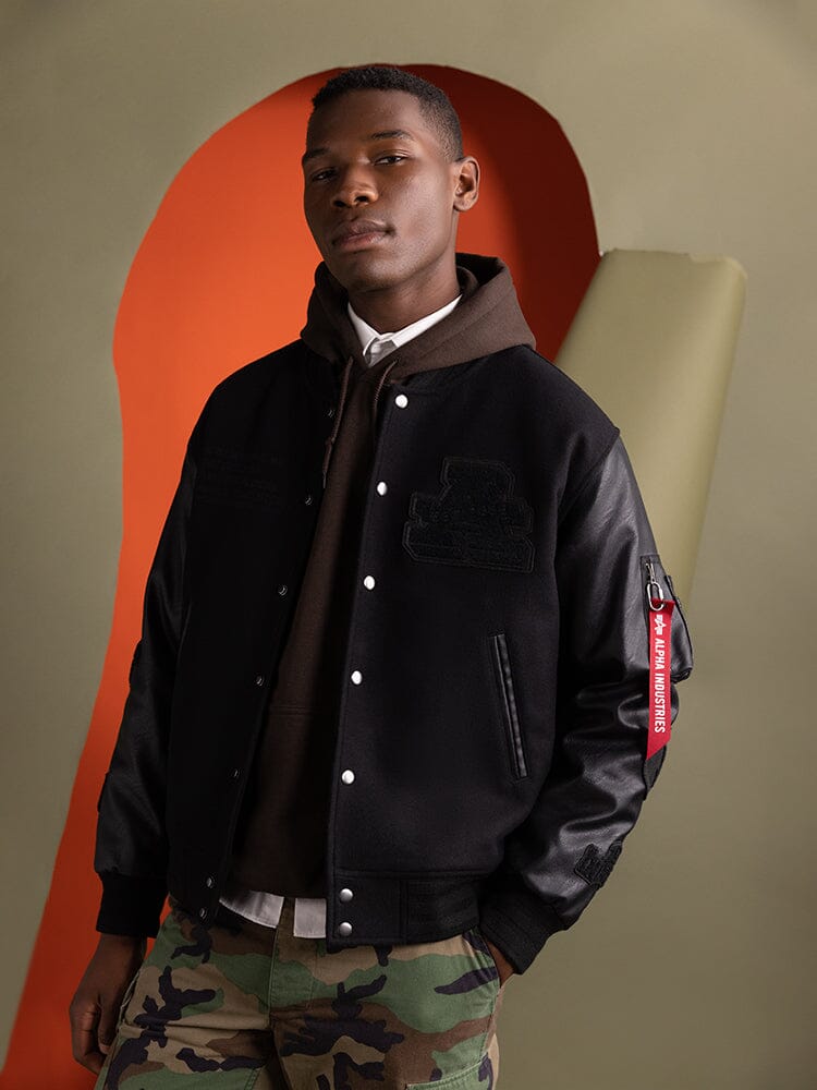 An individual wearing an Alpha Industries Faux Leather MA-1 Varsity Bomber Jacket in black.