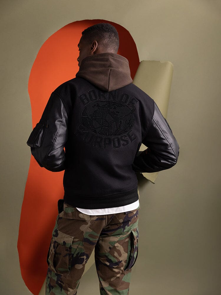 A person wearing the Faux Leather MA-1 Varsity Bomber Jacket in black by Alpha Industries, featuring "Born of Purpose" on the back.