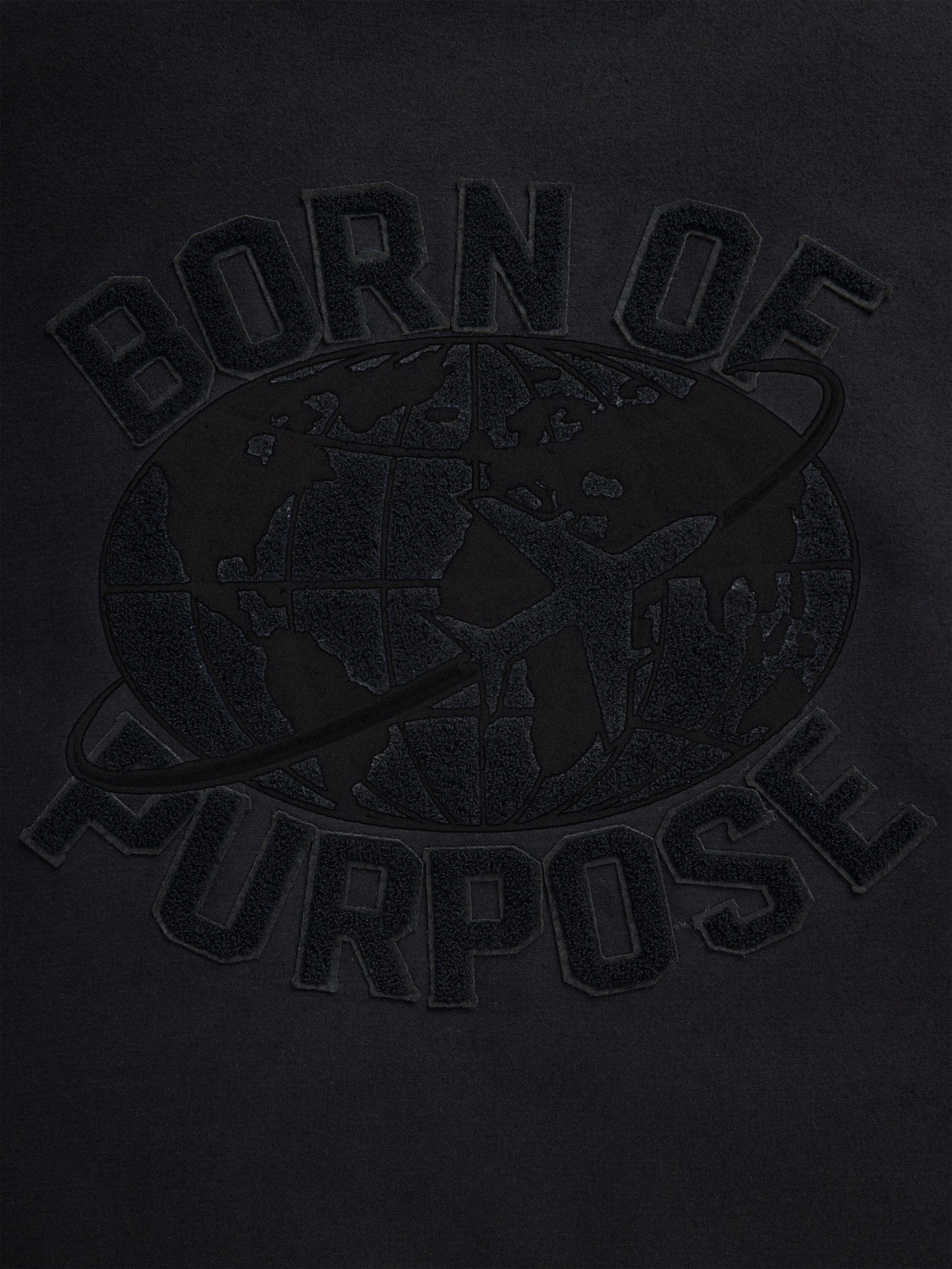 Close-up of the Faux Leather MA-1 Varsity Bomber Jacket from Alpha Industries features a black fabric design with "Born of Purpose" text and a world map graphic overlaid, accented by an orbit line circling the globe.