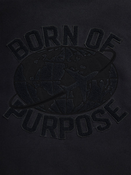 Close-up of the Faux Leather MA-1 Varsity Bomber Jacket from Alpha Industries features a black fabric design with "Born of Purpose" text and a world map graphic overlaid, accented by an orbit line circling the globe.