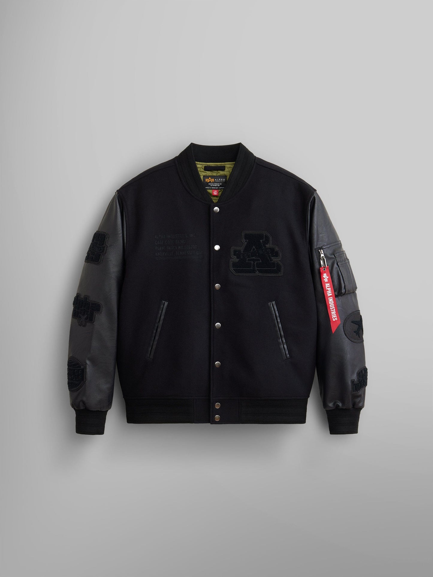 The Faux Leather MA-1 Varsity Bomber Jacket by Alpha Industries showcases a black wool blend body with dark gray faux leather sleeves, adorned with embroidered patches, front buttons, and a distinctive red zip tag on the sleeve.