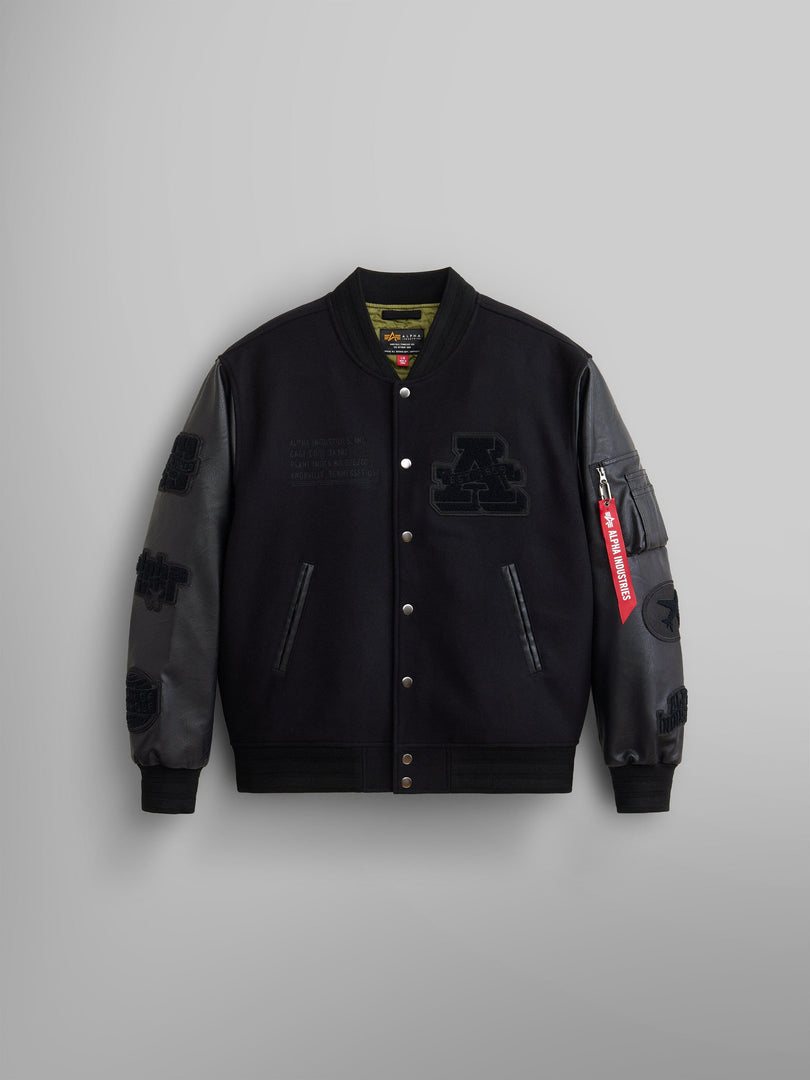 The Faux Leather MA-1 Varsity Bomber Jacket by Alpha Industries showcases a black wool blend body with dark gray faux leather sleeves, adorned with embroidered patches, front buttons, and a distinctive red zip tag on the sleeve.