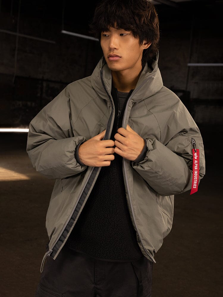 An individual in an Alpha Industries Insulated Hooded Parka with responsible down fill stands indoors, adjusting the zipper of their water-resistant coat.