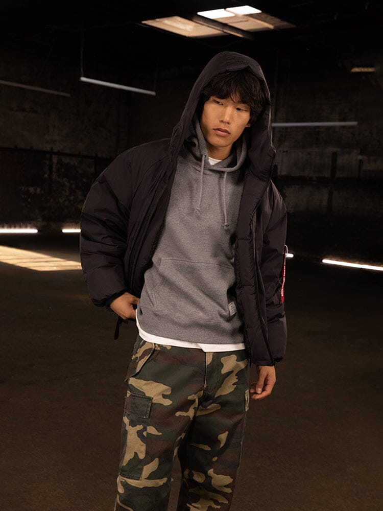 Clad in the Alpha Industries Insulated Hooded Parka, a person stands stylishly in camo pants, effortlessly showcasing the versatility of their water-resistant attire.