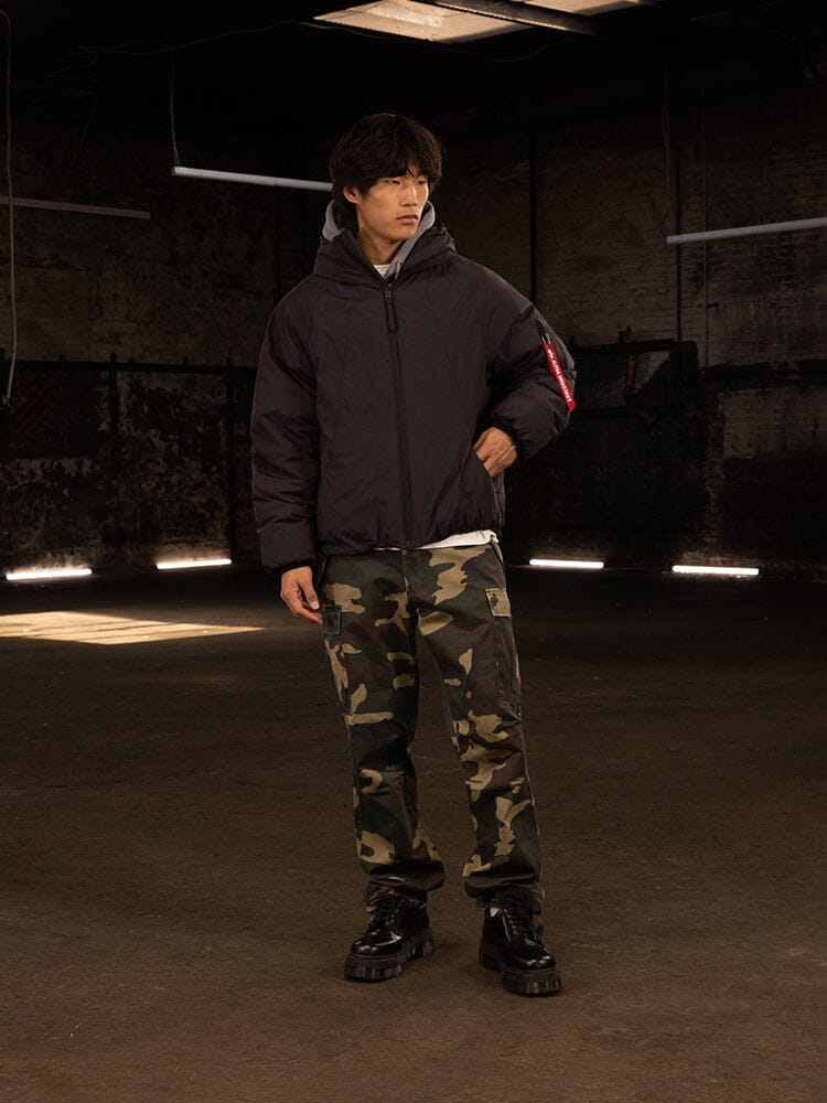 A person wearing an Alpha Industries Insulated Hooded Parka and camo pants stands in a dimly lit room.