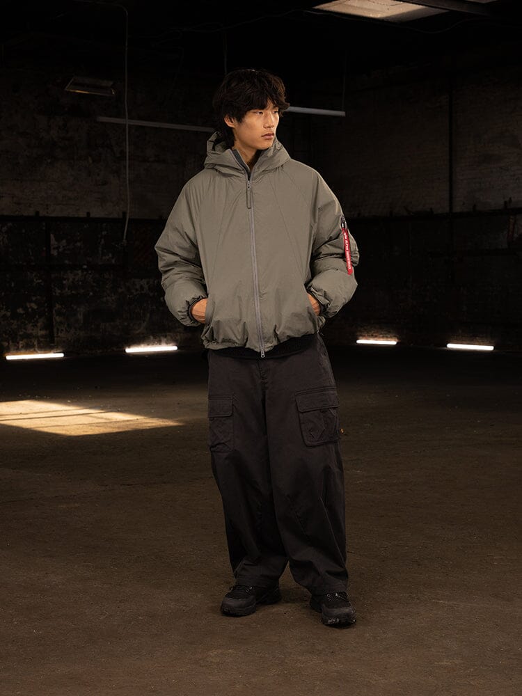 An individual stands wearing the Alpha Industries Insulated Hooded Parka with responsible down fill and black cargo pants, gazing to the side.