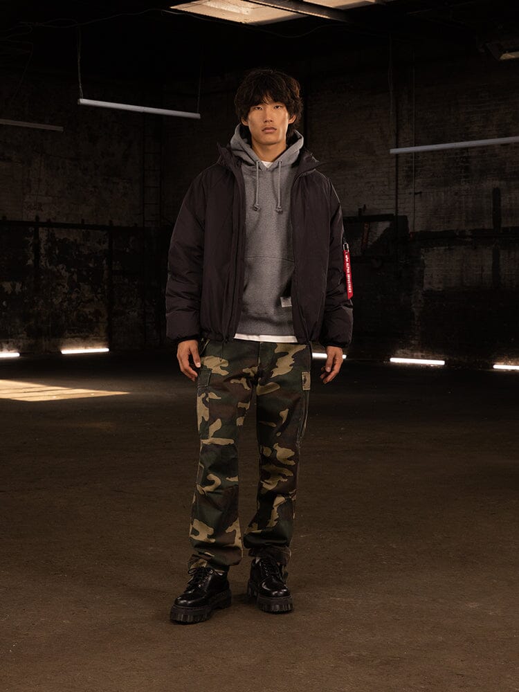 A person stands wearing an Alpha Industries Insulated Hooded Parka with responsible down fill, a gray hoodie, camouflage pants, and water-resistant black boots.