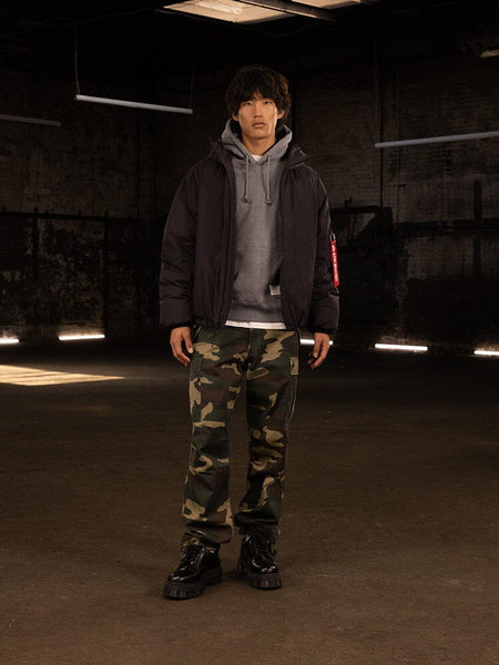 A person stands wearing an Alpha Industries Insulated Hooded Parka with responsible down fill, a gray hoodie, camouflage pants, and water-resistant black boots.