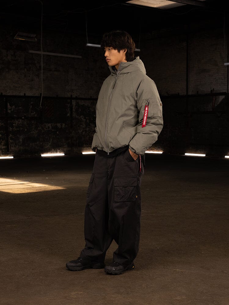 A person wearing an Alpha Industries Insulated Hooded Parka, paired with black cargo pants, stands in a dimly lit room; the parka's water-resistant fabric and responsible down fill offer comfort and sustainability.