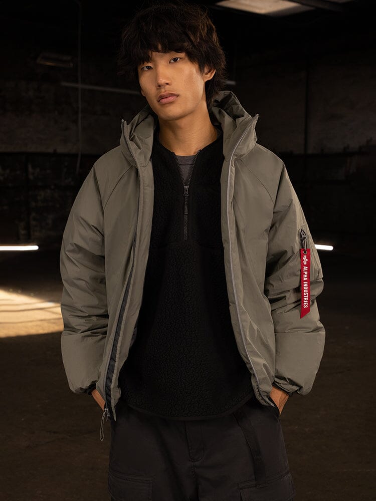 An individual wearing the Alpha Industries Insulated Hooded Parka, featuring a gray exterior with a distinctive red tag on the sleeve and filled with responsibly sourced down.