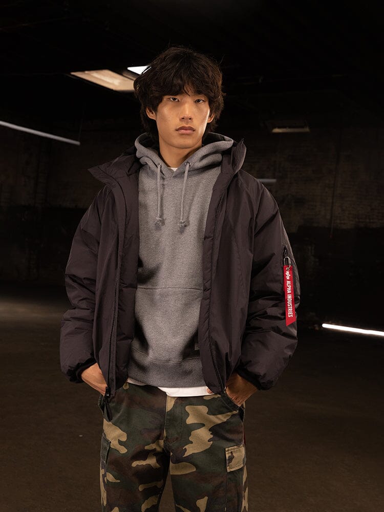 A person wearing the Insulated Hooded Parka by Alpha Industries, featuring a black puffer jacket with responsible down fill, along with camouflage pants.