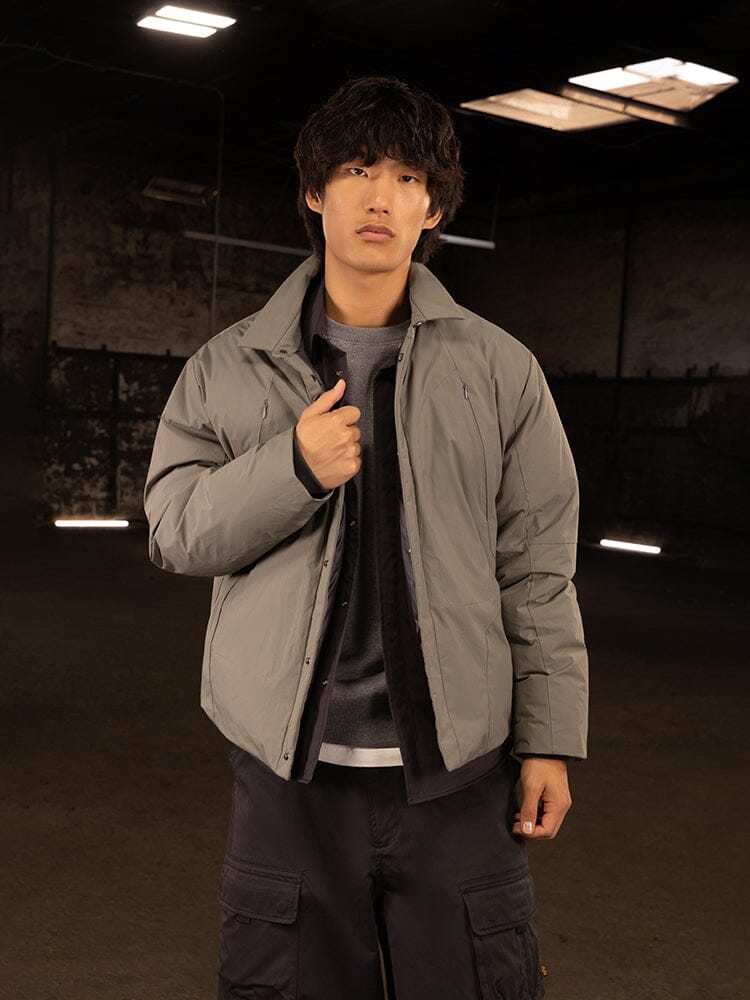 In a dimly lit industrial setting, a person exudes streetwear style by donning the gray Insulated Shirt Jacket from Alpha Industries over a dark shirt and black pants.