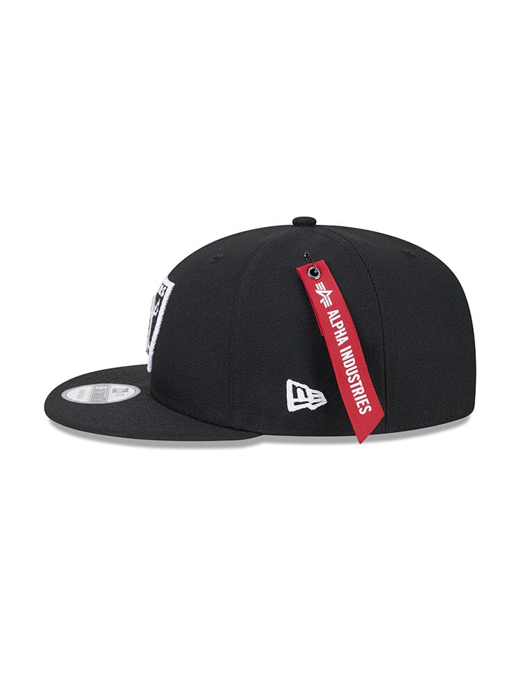 Black Las Vegas Raiders X Alpha X New Era 950 Cap with a flat brim, showcasing Alpha Industries' signature red tag at the back and a white emblem on the side.