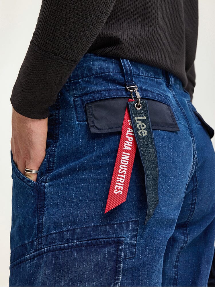 An individual wearing the Alpha X Lee ACU Pant by Alpha Industries, designed with blue ripstop cotton in a military style, and accented with a red Remove Before Flight tag keychain attached to the cargo pocket.