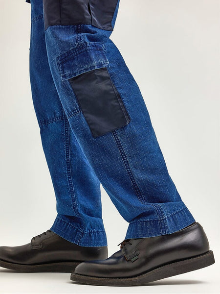 A close-up of someone wearing the Alpha X Lee ACU Pant from Alpha Industries, showcasing blue denim pants with cargo pockets and a distinctive black pocket patch paired with black dress shoes, all made from ripstop cotton.