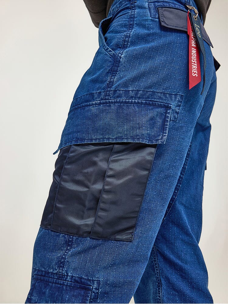 Close-up of a person wearing the Alpha X Lee ACU Pant by Alpha Industries, showcasing blue cargo pants made from ripstop cotton with black patchwork in a military design and featuring a red tag on one of the cargo pockets.