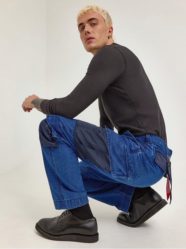 A person with short blond hair crouches, dressed in a black long-sleeve shirt and the Alpha X Lee ACU Pant from Alpha Industries. The pants are blue ripstop cotton cargo pants featuring dark patches and cargo pockets, paired with black shoes.