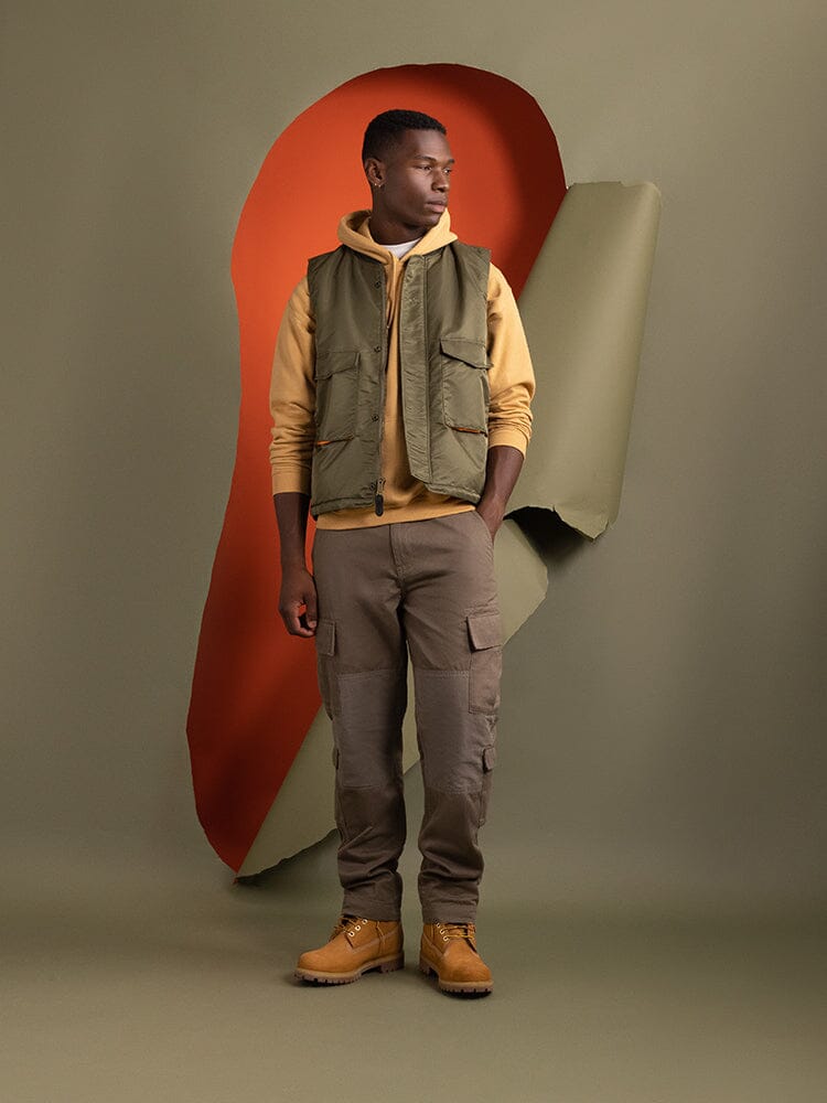 A person wearing Alpha Industries' MA-1 Mod Vest in OG-107 Green, complemented by a yellow hoodie and cargo pants.