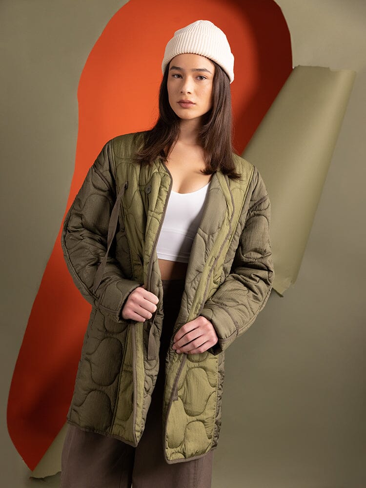 A woman sports the Women's Mixed Media Quilted Long Liner in OG-107 Green by Alpha Industries, and a white crop top, effortlessly embracing the layering trend.