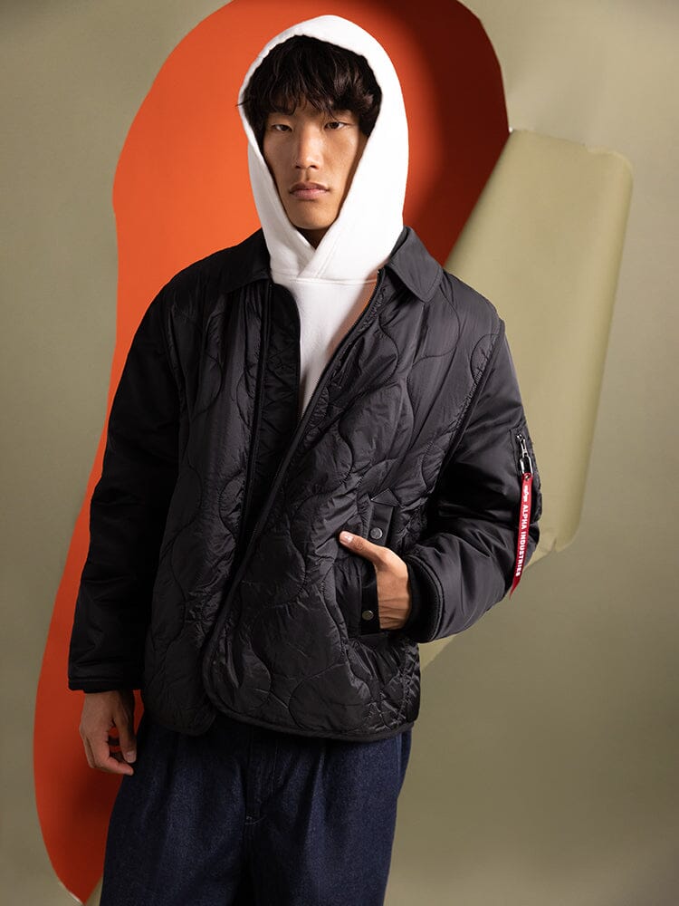 A person wears a white hoodie and the Mixed Media Quilted Utility Jacket in black by Alpha Industries.