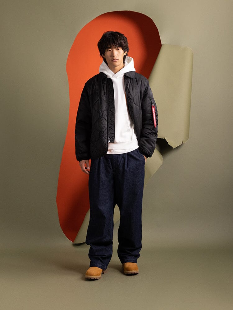 A person stands showcasing the Mixed Media Quilted Utility Jacket in black by Alpha Industries, layered over a white hoodie and paired with blue pants.