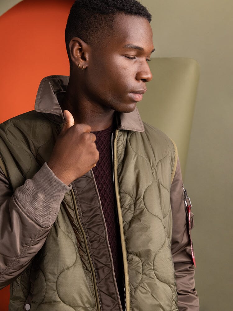 An individual wearing the Mixed Media Quilted Utility Jacket in coyote brown by Alpha Industries stands with a thoughtful sideways glance.