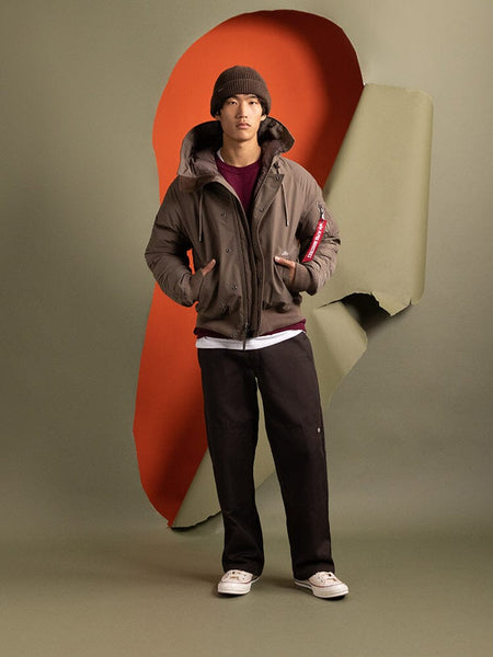 A person wearing an Alpha Industries N-2B Altitude Parka, a water-resistant brown jacket, dark pants, and a beanie.