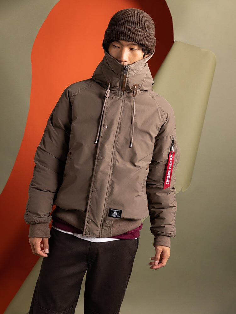 A person wearing a brown Alpha Industries N-2B Altitude Parka, paired with an adjustable split hood, beanie, and brown pants.