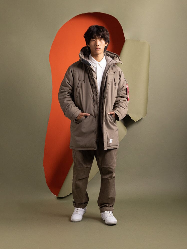 A person stands dressed confidently in a brown N-3B Altitude Mod Parka from Alpha Industries.