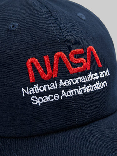 This close-up showcases the NASA Worm Logo Cap by Alpha Industries. The cap is a navy blue baseball style with the iconic NASA worm logo embroidered in striking red and white.