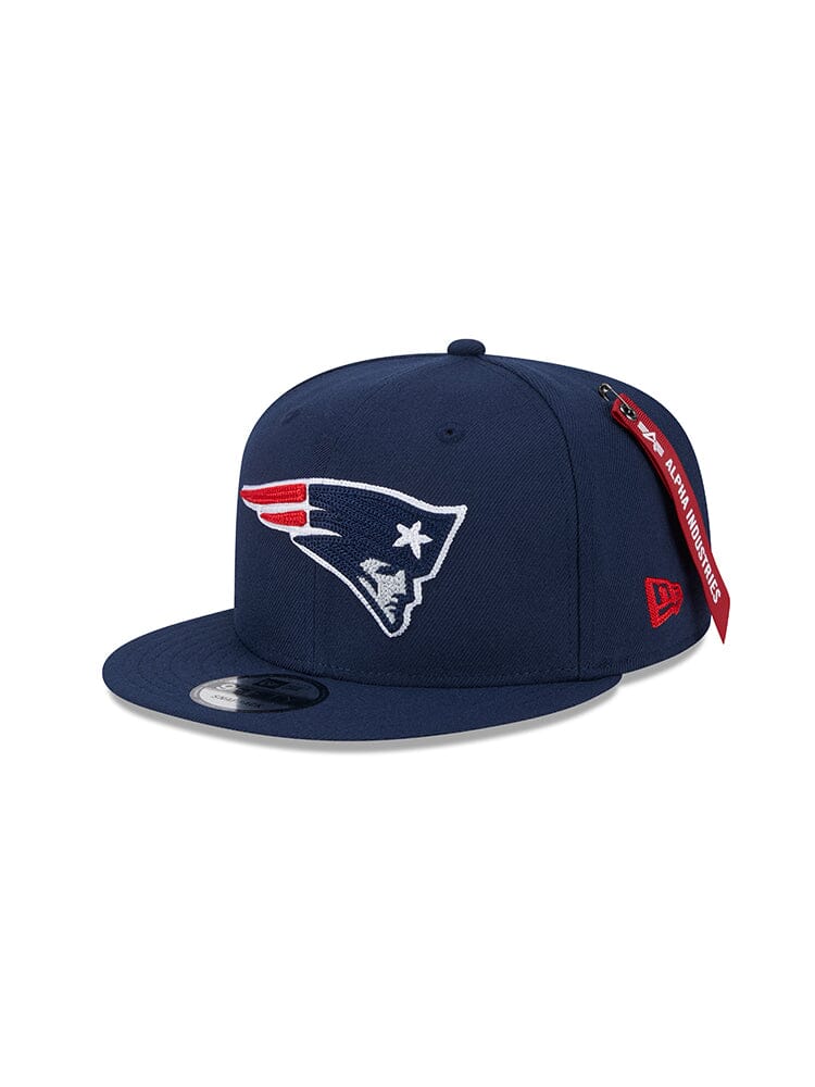 This New England Patriots x Alpha x New Era 950 Cap from Alpha Industries features a dark blue design with a red and white stylized face logo, complete with side tags showcasing the NFL and 9FORTY logos.