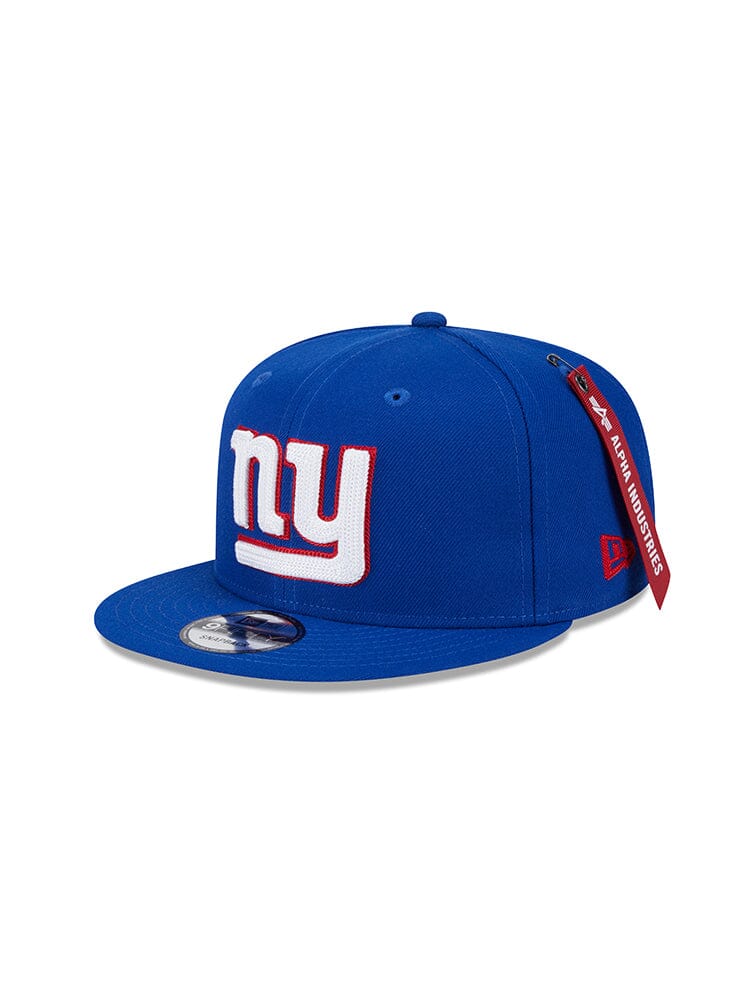 Blue snapback baseball cap from the New York Giants x Alpha Industries x New Era 950, featuring the iconic NY team logo in white and red on the front. A red tag on the side highlights Alpha Industries® style.