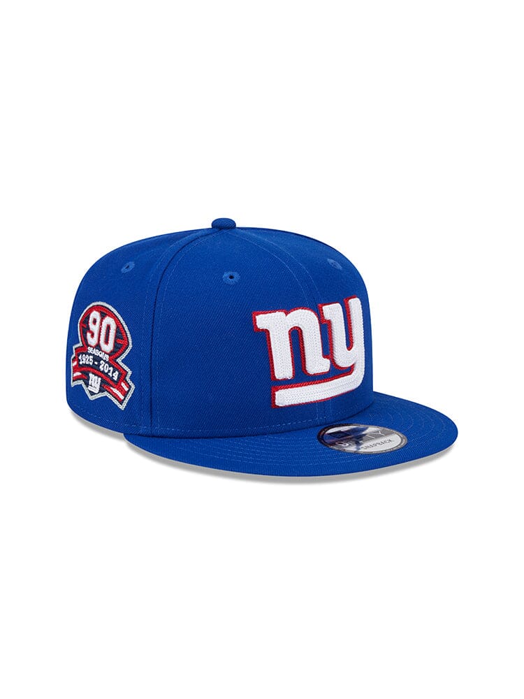 Snapback blue baseball cap crafted with signature Alpha Industries® quality, featuring the iconic NY team logo on the front and a 90 years 1925-2014 commemorative patch on the side, named the New York Giants X Alpha X New Era 950 Cap.