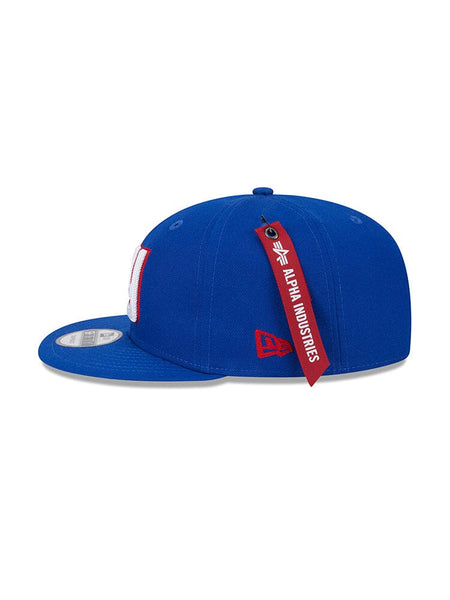 Introducing the New York Giants x Alpha x New Era 950 Cap, a sleek blue snapback baseball hat featuring a flat brim with the iconic red and white NY team logo on the front and an authentic red Alpha Industries® tag on the side.