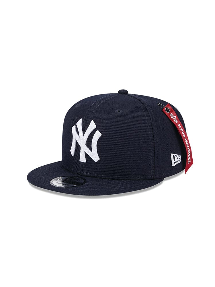 The New York Yankees x Alpha x New Era 950 Cap (Navy) by Alpha Industries features a flat brim and the iconic white NY team logo on the front. A distinctive red tag on the side adds a touch of flair to this navy cap.