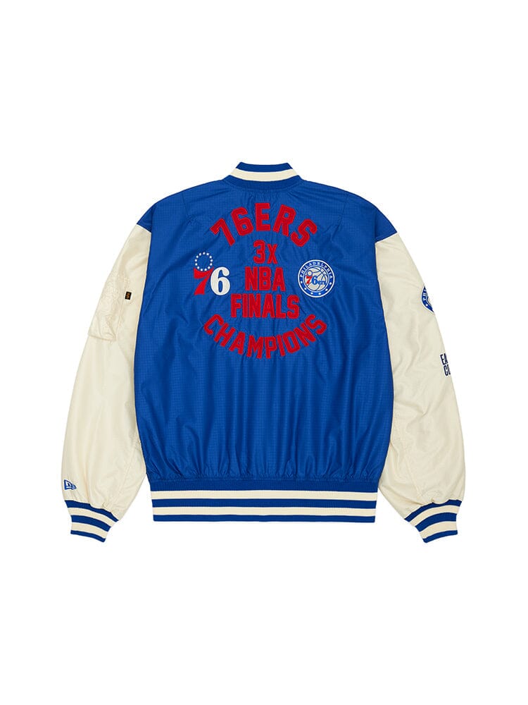 The Philadelphia 76ers x Alpha x New Era L-2B Bomber Jacket by Alpha Industries and NE-NBA is blue and white, featuring the text "76ers 3x NBA Finals Champions" with team patches on the back.