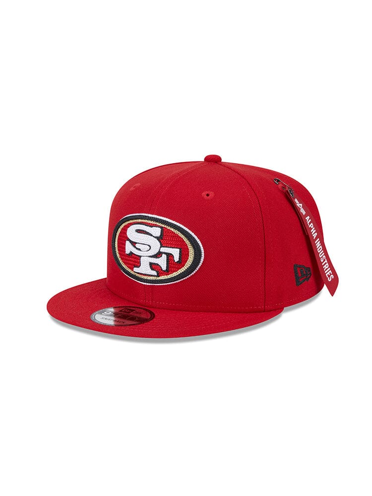 Introducing the San Francisco 49ers x Alpha Industries x New Era 950 Cap by Alpha Industries, featuring a prominent San Francisco 49ers logo on the front and a unique AAPE fabric tag on the side. This red cap embodies the classic New Era Snapback style, providing both comfort and an authentic athletic appearance.