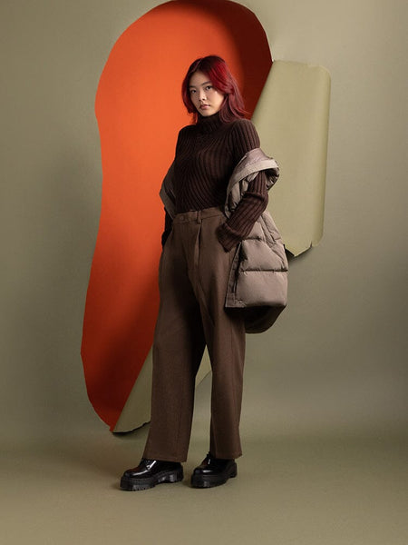 A person with red hair stands wearing a brown sweater and pants, with an Alpha Industries Sierra Convertible Jacket W draped over one shoulder.